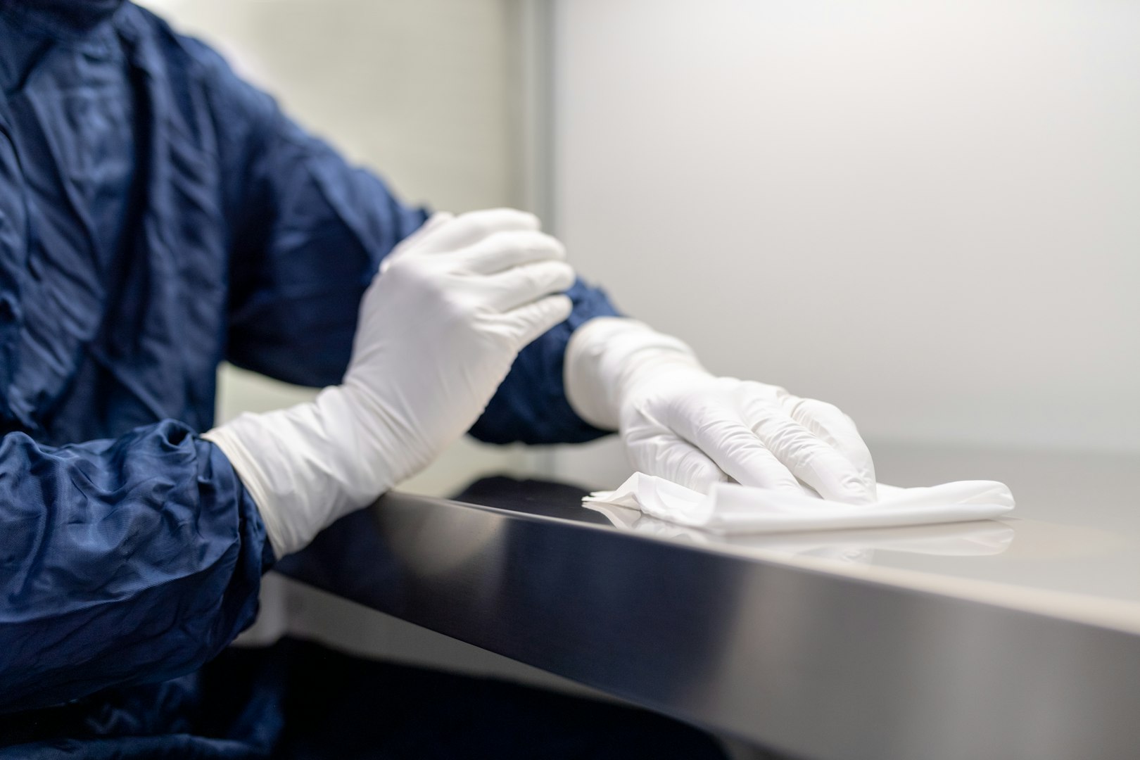 A person wearing a white shirt and blue overalls, with yellow rubber gloves, is cleaning a glass window with a cloth. In the background, two other individuals are engaged in cleaning tasks in an office environment.
